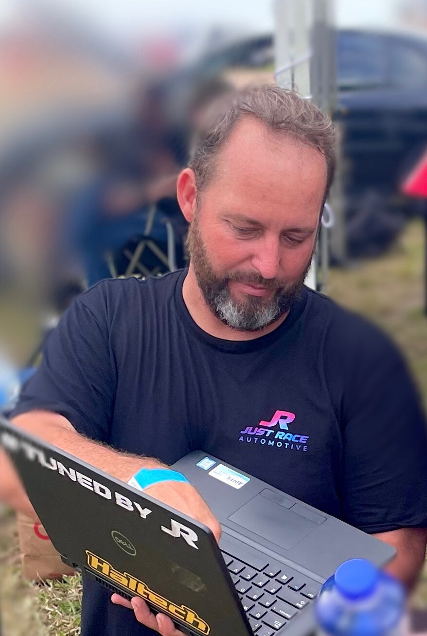 Caucasian man with beard wears a black shirt featuring “JR Just Race Automotive” logo in bright pink, blue and purple. He’s tuning a car, holding a laptop featuring the hashtag “Tuned by JR”. JR is an experienced car tuner who specialises in tuning drag cars, off-street racing and no-prep events in Australia.